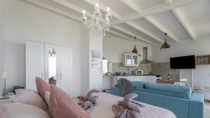 Une chambre à coucher avec un lit, des oreillers, un lustre, un canapé, une télévision et, au fond, une petite cuisine.