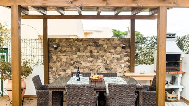 A covered outdoor dining area with wicker furniture, a stone wall, and a grill. - Holiday Home 'Laguna Formosa' with Private Terrace, Wi-Fi, SPA/Jacuzzi & Air Conditioning (Faro)