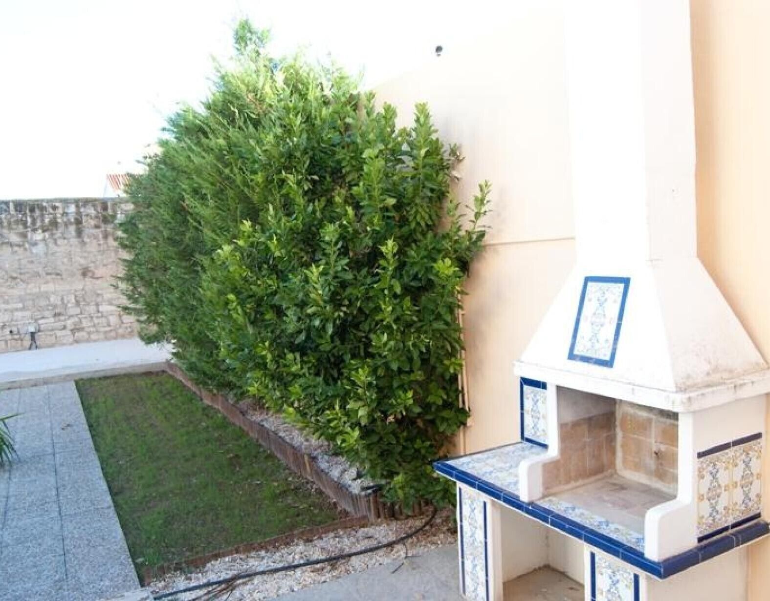 Un patio con muro de piedra, un pequeño césped y una cocina exterior blanca con detalles azules.