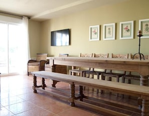 A dining area with a long table, benches, wicker chairs, and framed botanical prints on the wall.