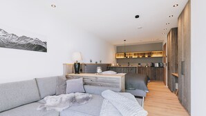 Ein modernes Wohnzimmer mit einem grauen Sofa, einem kleinen runden Tisch und einer Küchenzeile im Hintergrund.