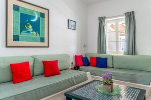 Een woonkamer met twee groene banken, rode en blauwe kussens, een ingelijst schilderij, een raam met gordijnen en een bijzettafeltje met een plant.