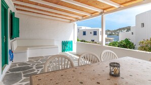 Un área de estar al aire libre con techo, una mesa y sillas, vista al mar y edificios blancos en el fondo.