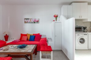 A compact living space with a sofa, a small dining table, a washing machine, and a kitchenette.