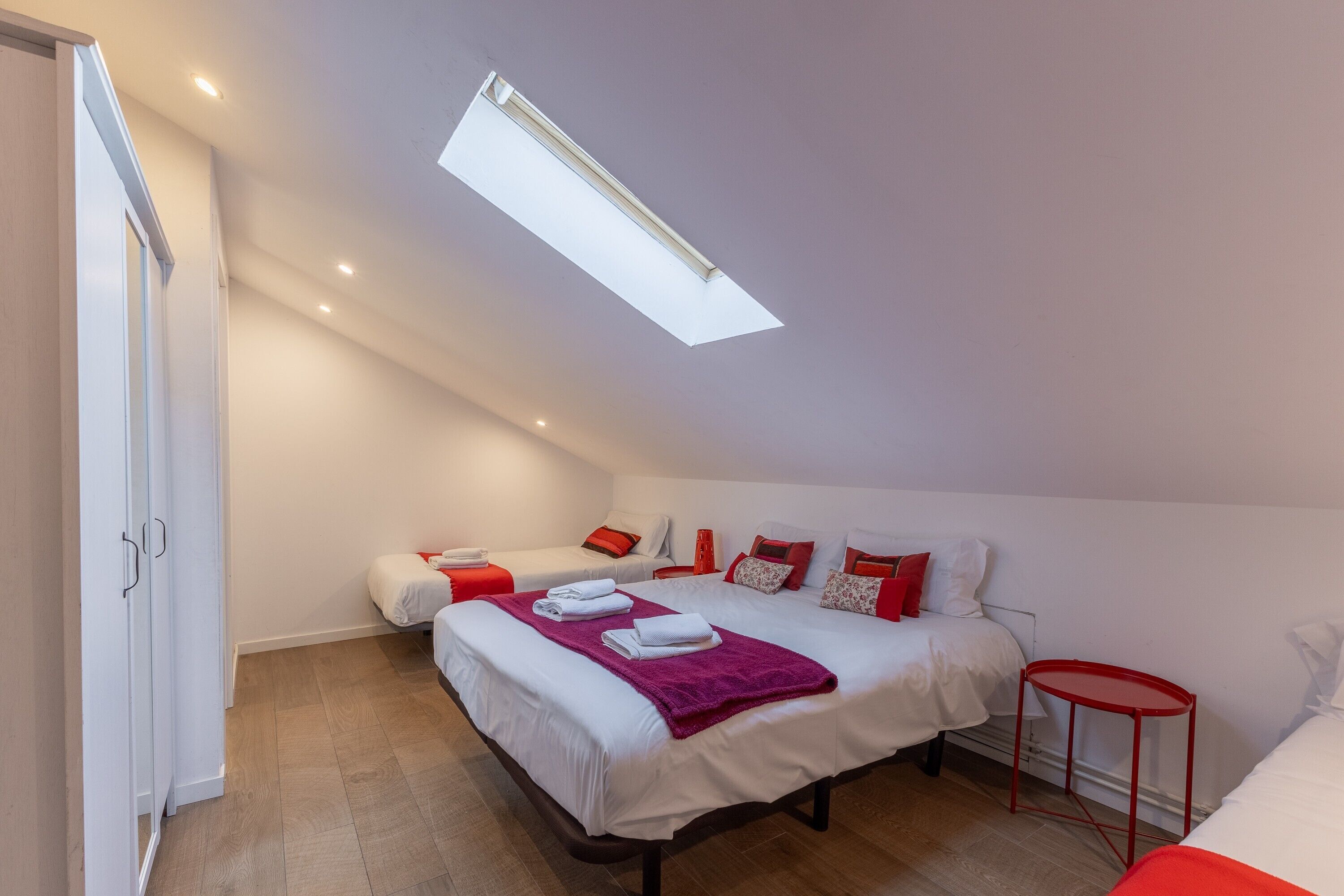 A bedroom with two beds, a skylight, a red side table, and a wardrobe.