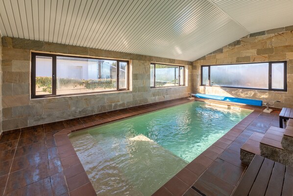 Una piscina con paredes de piedra y ventanas amplias, techo de listones de madera y una zona con bancos.
