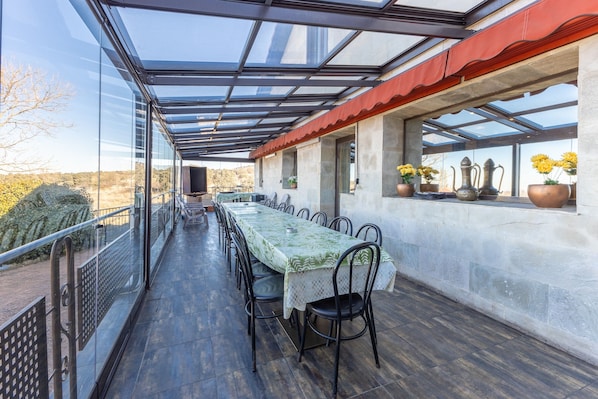 Un área de comedor al aire libre techada, con una mesa larga, sillas, un cerramiento de vidrio y plantas en macetas.