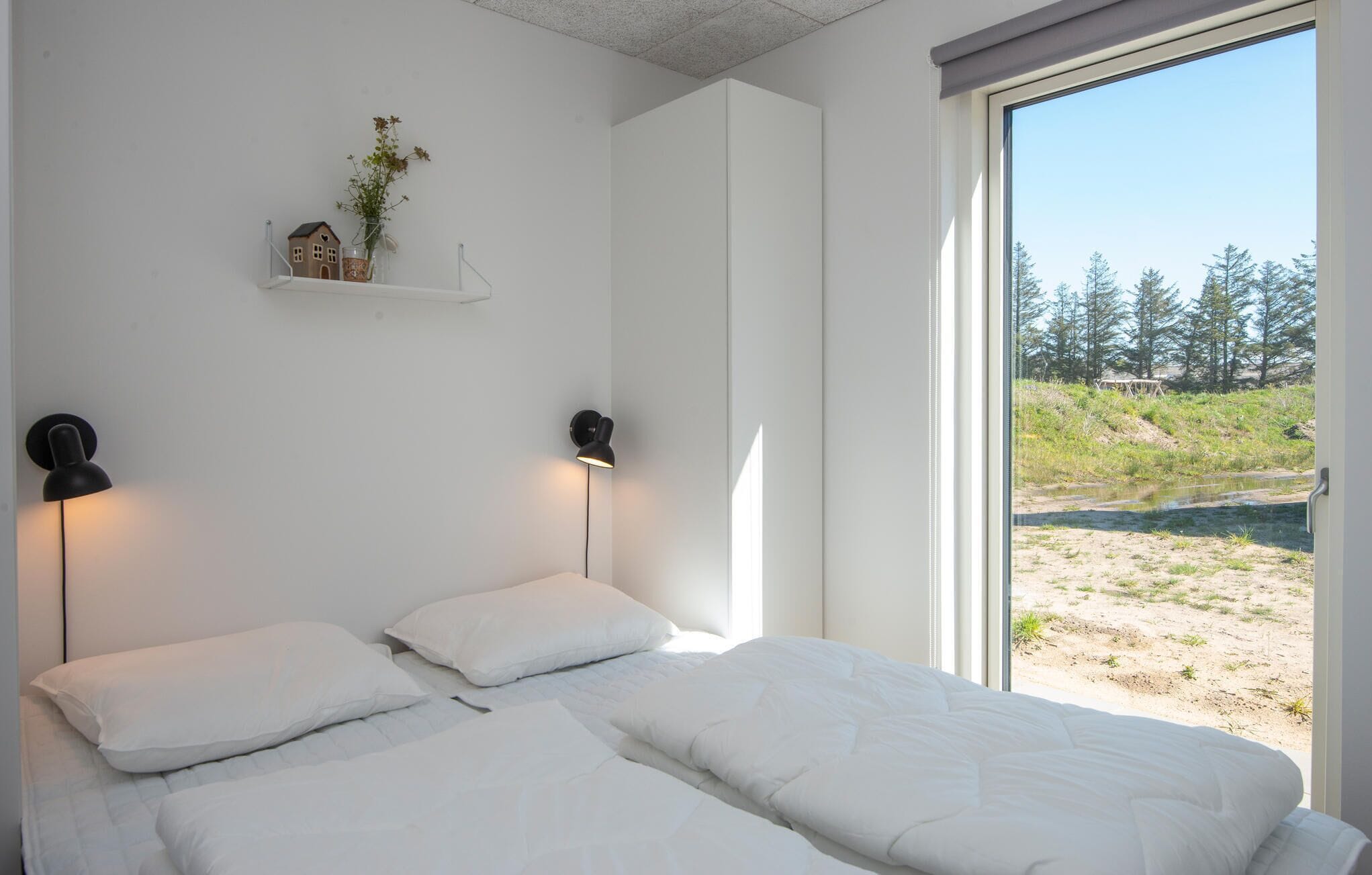 Ein Schlafzimmer mit zwei Betten, einer kleinen Ablage mit einem Hausmodell und Blumen, zwei Wandleuchten und einem großen Fenster mit Blick auf einen Strand und Bäume.