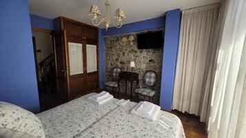 A bedroom with a bed, a chandelier, a stone wall, a television, and a window with curtains.