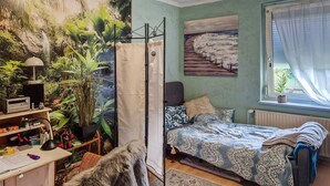 A bedroom with a bed, a window with curtains, a desk with a computer, and a wall with a painting.