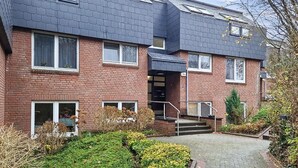 A two-story brick building with a dark roof, a central entrance with steps, and a small garden area with shrubs and a tree.