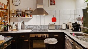 A kitchen with a stove, oven, and various appliances. There are shelves with jars and bottles, and a clock on the wall.