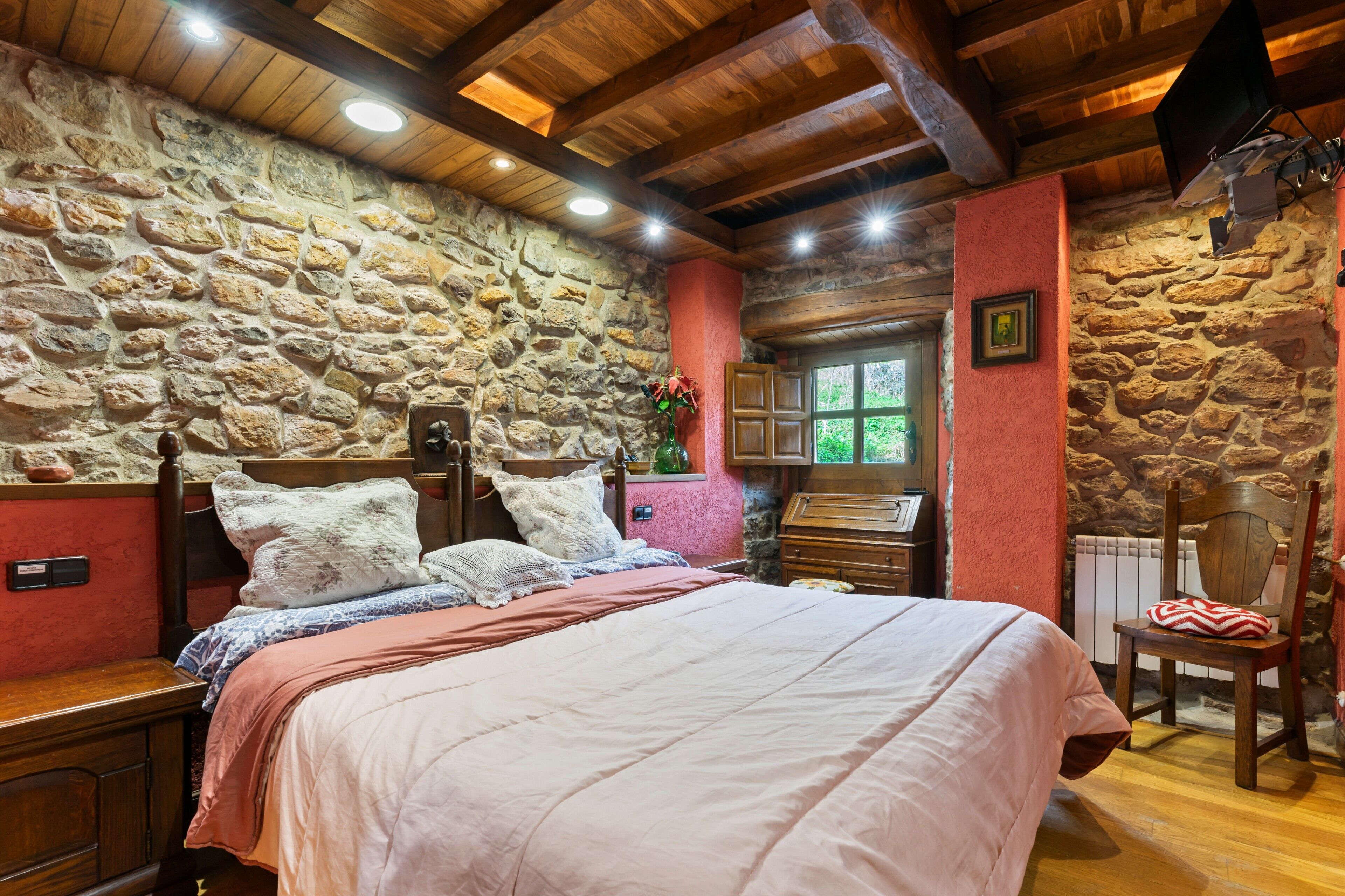 Een slaapkamer met een stenen muur, houten balken in het plafond, een bed met kussens, een dressoir en een stoel.