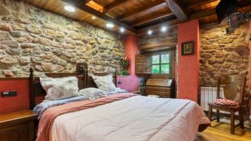 Een slaapkamer met een stenen muur, houten balken in het plafond, een bed met kussens, een dressoir en een stoel.