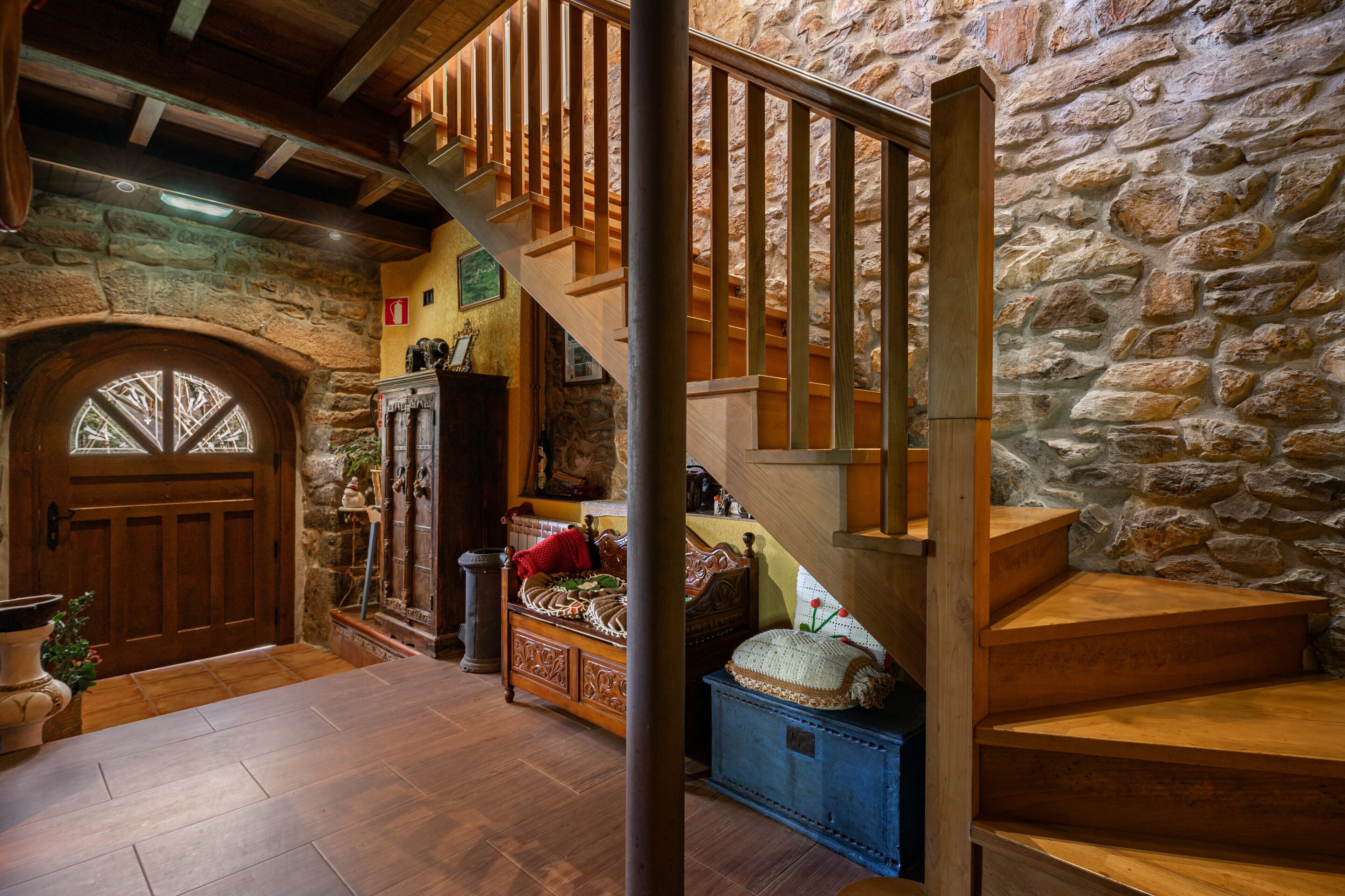 Un escalier en bois menant à un niveau supérieur dont les murs sont en pierre, une porte en bois avec un vitrage, et un lit recouvert d’une couverture rouge.