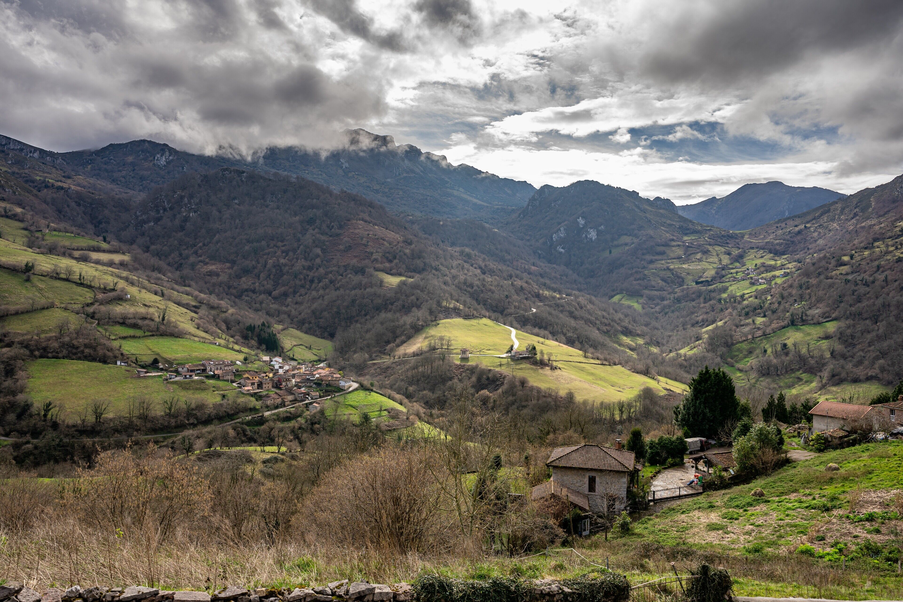 Un paisaje montañoso con un pequeño pueblo, muros de piedra y un camino sinuoso.