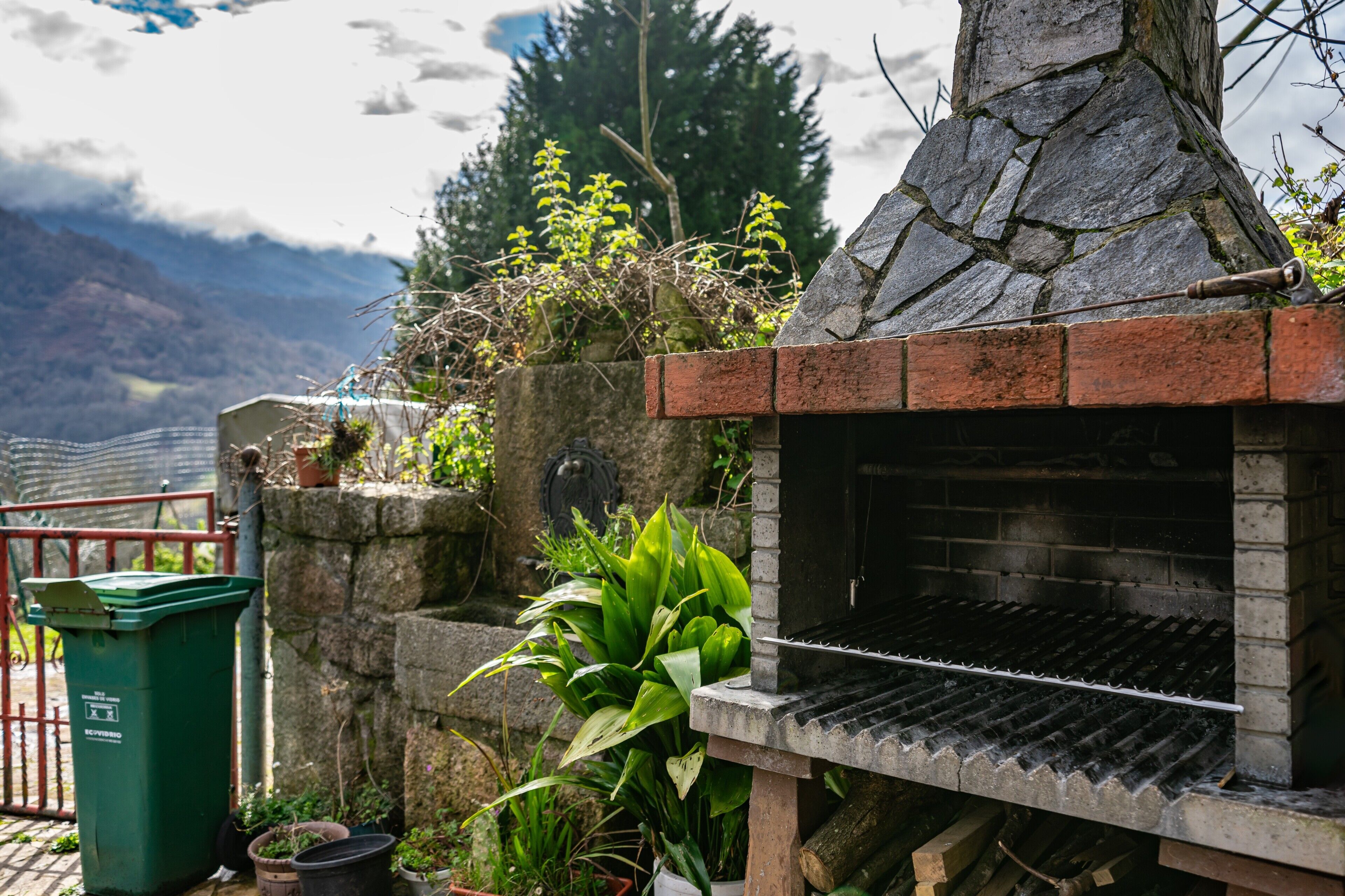 A stone-built outdoor grill with a brick base, surrounded by potted plants and a green trash bin.