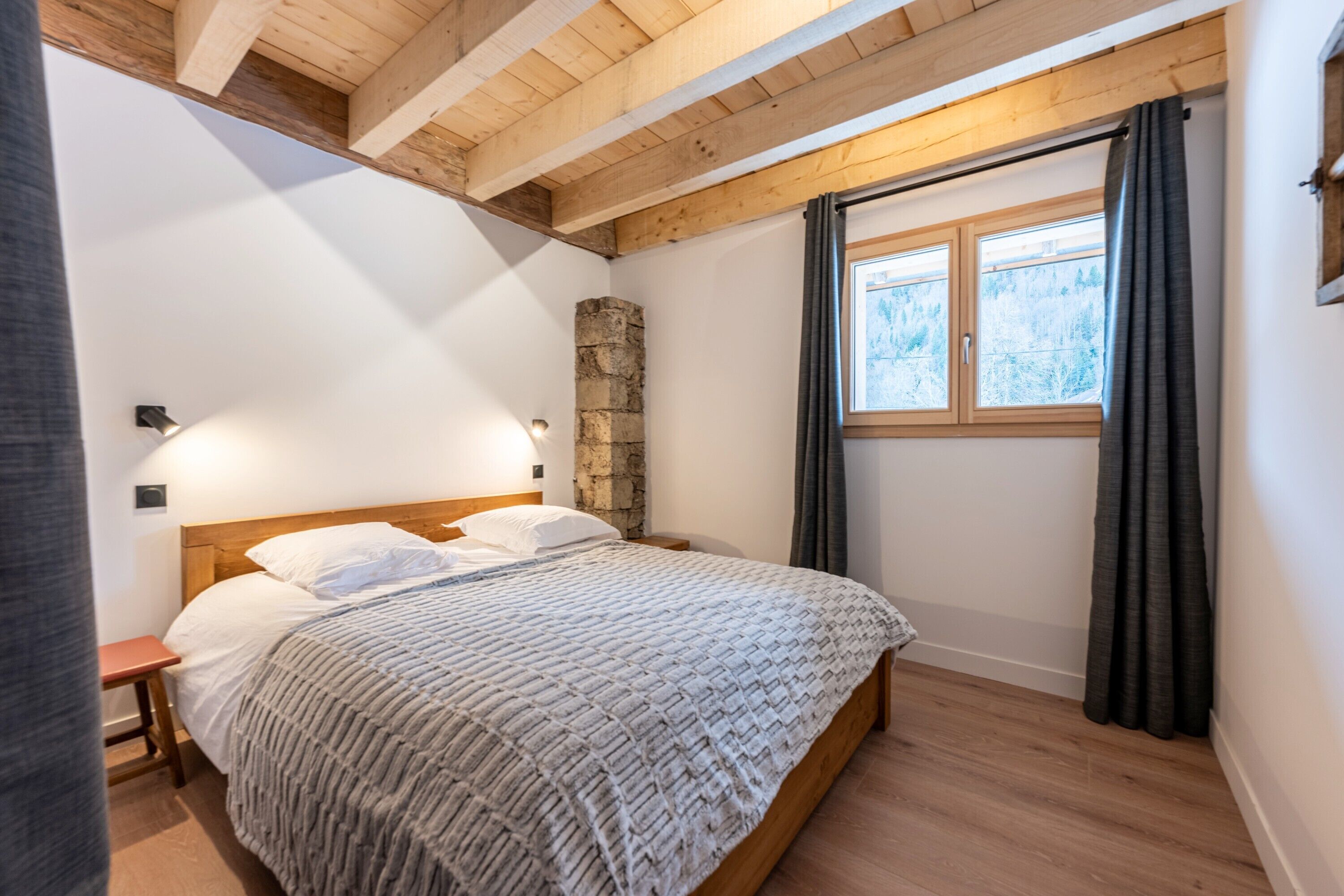 A bedroom with a wooden headboard, a stone accent wall, a window with curtains, and a bedside table.