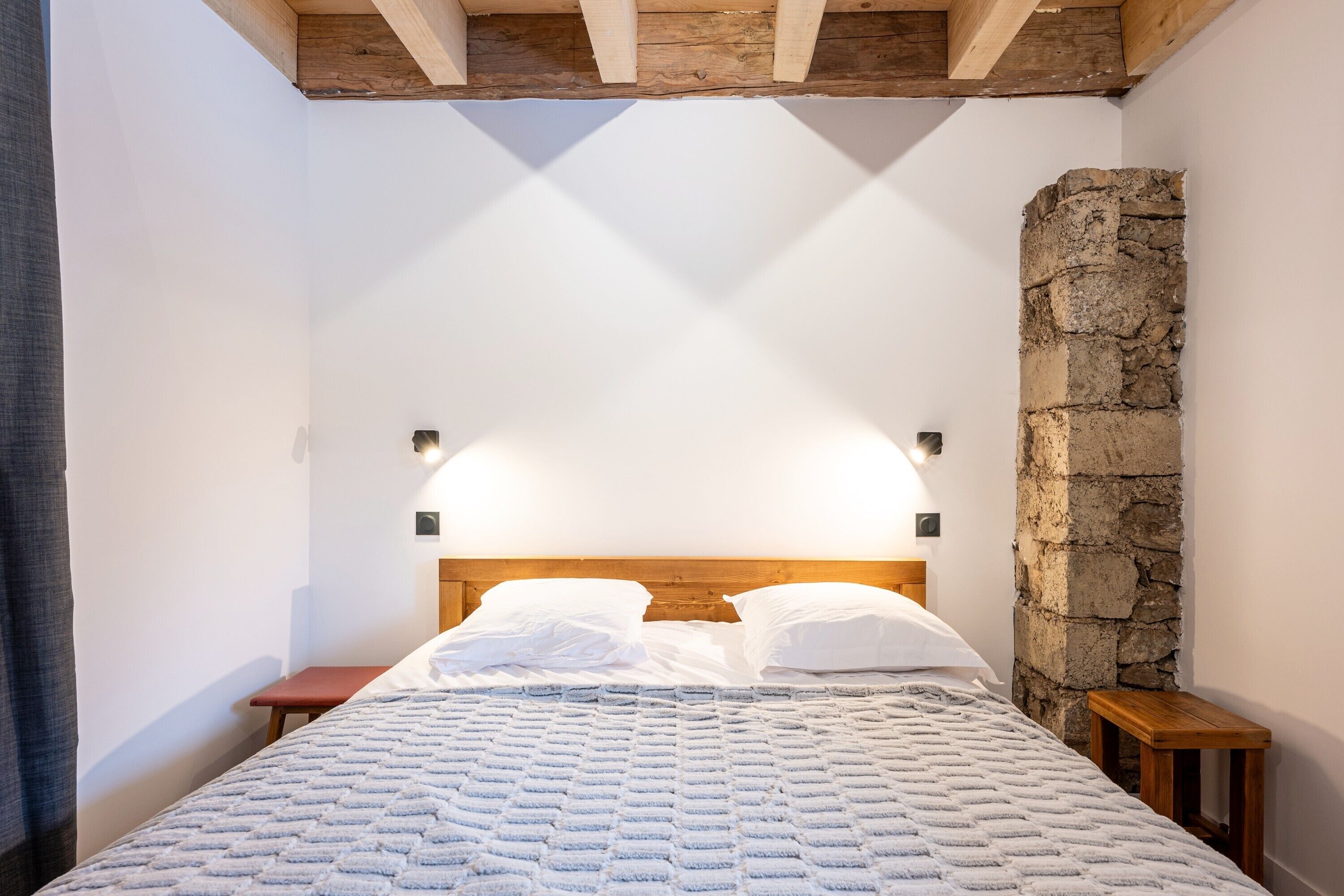 Une chambre  à coucher avec une tête de lit en bois, un mur en pierre et un lit recouvert de linge de lit blanc.