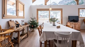 A dining area with a long table, wooden chairs, a small Christmas tree, and a television on a wooden cabinet.