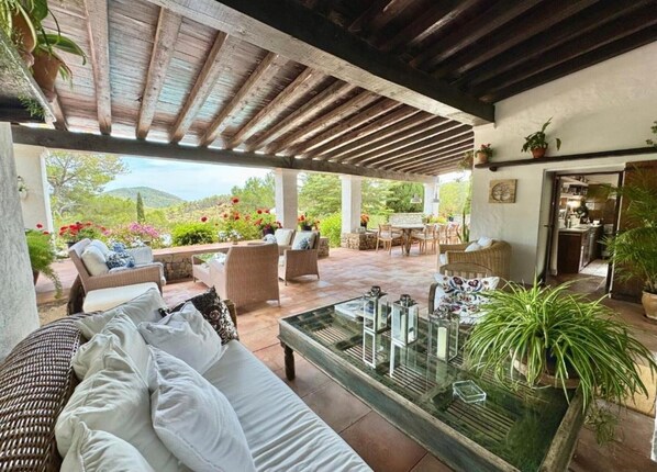 A covered outdoor seating area with wicker furniture, a glass-top table, and potted plants.