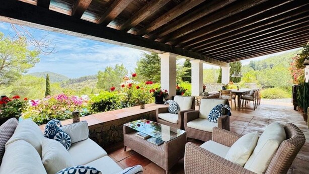 An outdoor patio area with wicker furniture, a glass-top table, and a view of a garden with flowers and trees. - Villa 'Ses Casetes Ibiza' with Mountain View, Wi-Fi and Air Conditioning (Illes Balears)