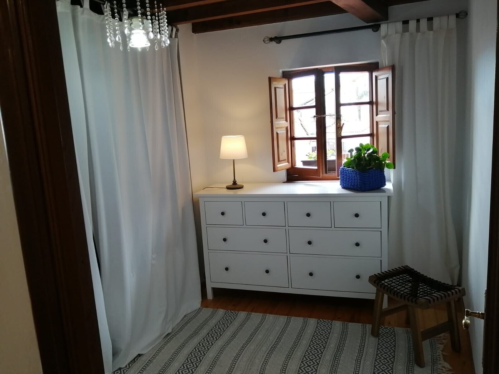 Une pièce avec une commode blanche, une lampe, une fenêtre avec des rideaux, un petit tabouret et un tapis à motifs.