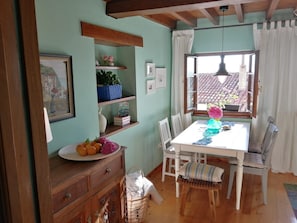 A dining area with a table, chairs, a window with curtains, a wooden cabinet, and shelves with decorative items. - Holiday Home 'El Canto De Las Gaviotas' with Sea View, Balcony and Wi-Fi (Lastres)