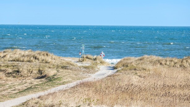 Een kustpad dat naar de zee leidt, met een reddingsboei en een bord dat een strand aangeeft.