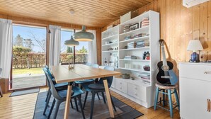Een gezellige kamer met een houten plafond, een eettafel met stoelen, een boekenplank, een gitaar en een lamp.