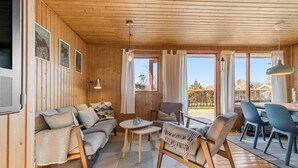 Een knus houten interieur met een bank, stoelen, een eettafel en uitzicht op de omgeving.