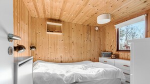 Een slaapkamer met een houten plafond, een bed met wit beddengoed, een klein raam, een radio en een witte dressoir.