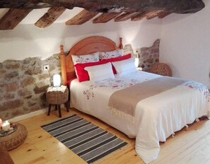 A bedroom with a wooden ceiling, stone walls, a bed with floral bedding, a bedside lamp, and a woven basket on the floor. - Country House 'Nela' with Mountain View, Private Terrace and Wi-Fi (Villaviciosa)