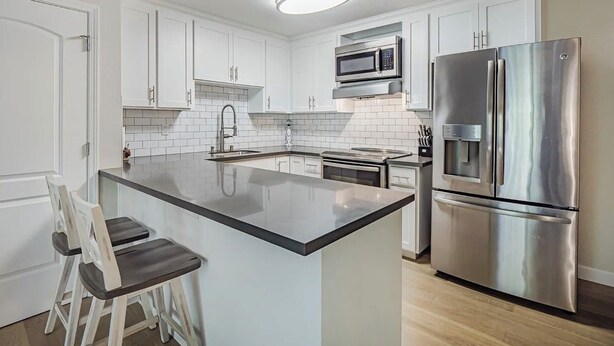 A modern kitchen with white cabinetry, a stainless steel refrigerator, a microwave, and a built-in island with a bar stool.