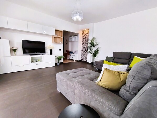 A modern living room with a grey sofa, a flat-screen TV, white cabinetry, and a decorative wall panel.