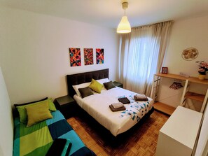 A bedroom with a bed, a bedside table, a desk, and a hanging light fixture.