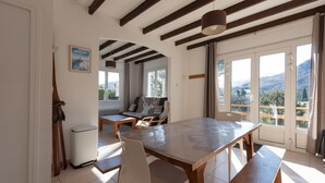 Un'area pranzo con tavolo e sedie in legno, una panca, un divano con cuscini, un quadro appeso e ampie finestre che offrono vista sull'esterno.