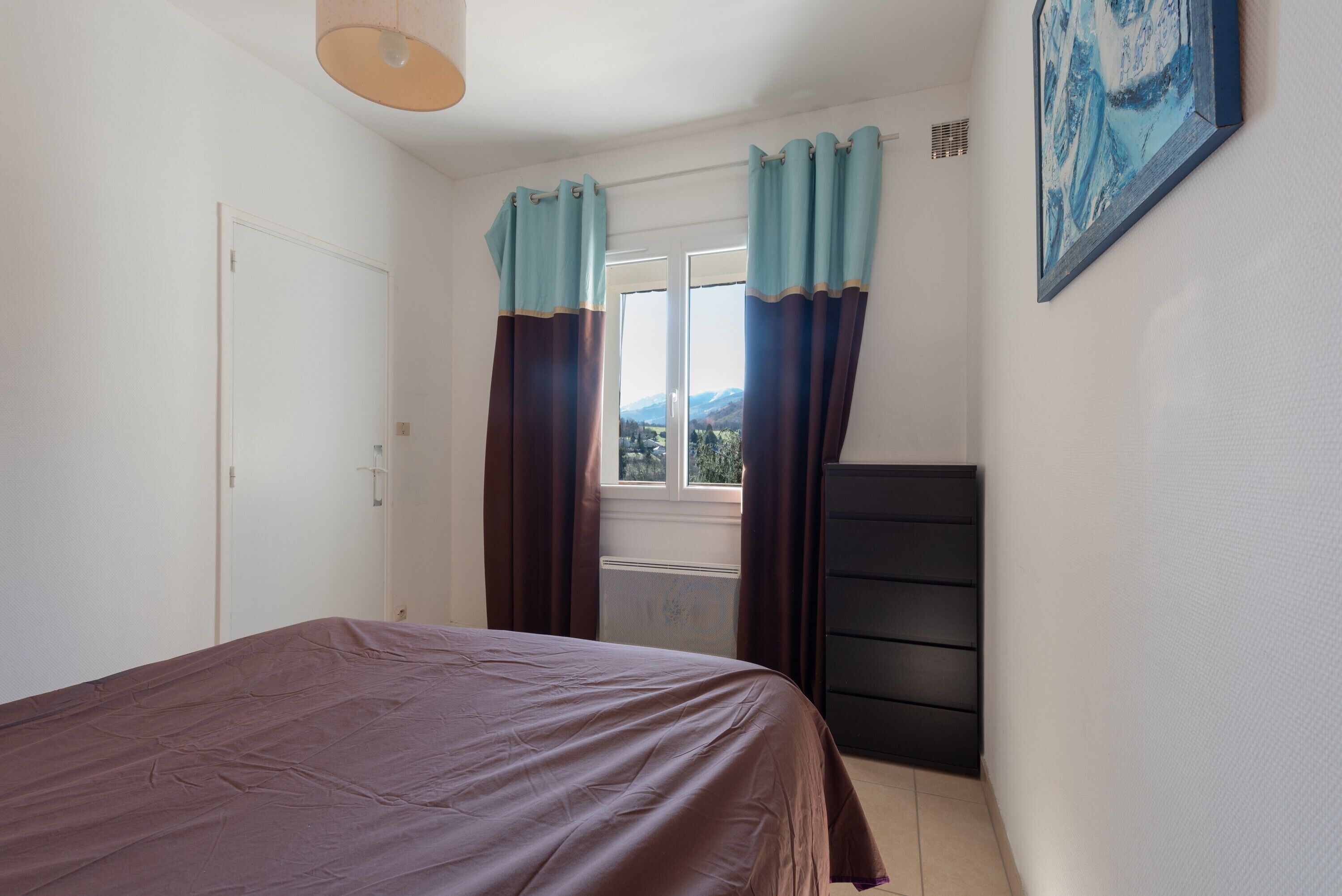 A bedroom with a bed, a dresser, a window with curtains, and a painting on the wall.