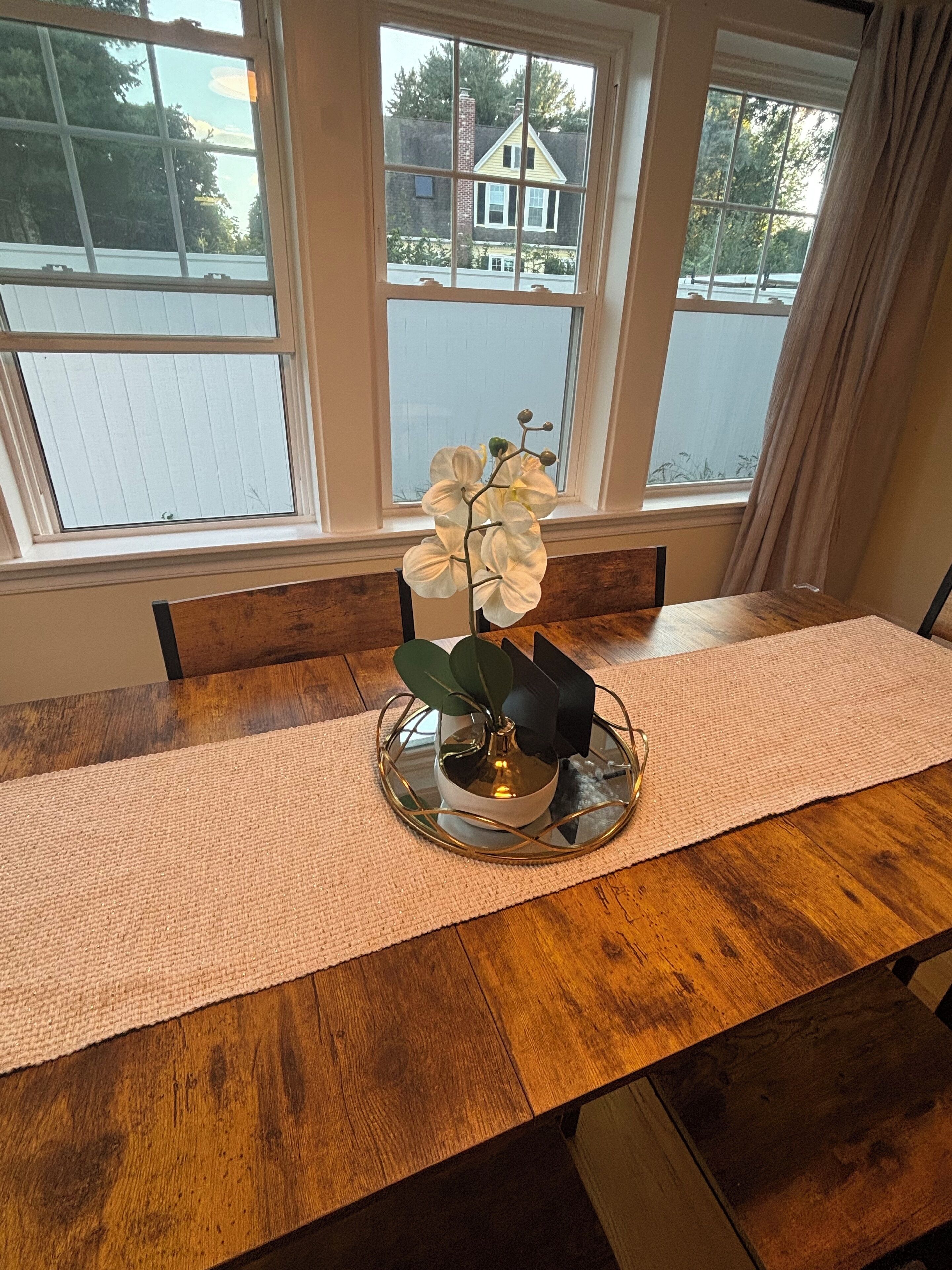 A dining table with a white orchid centerpiece, a woven placemat, and a view of a residential area through large windows.