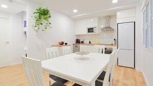 Une cuisine moderne avec des meubles blancs, un four à micro-ondes, une cuisinière et un réfrigérateur. On y trouve une table à manger avec des chaises et une plante suspendue.