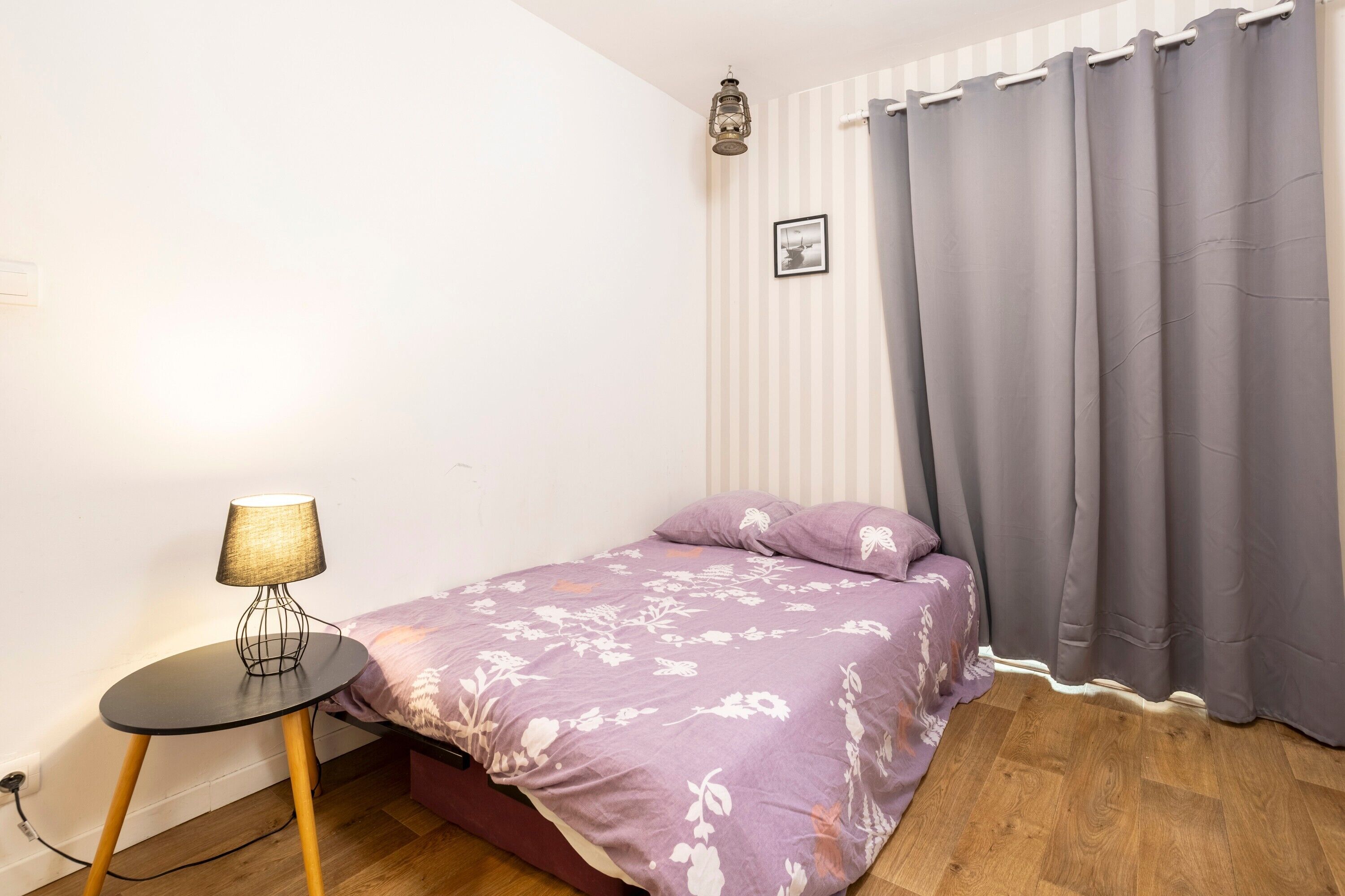 A bedroom with a bed, a small round table with a lamp, a picture on the wall, and a window with curtains.