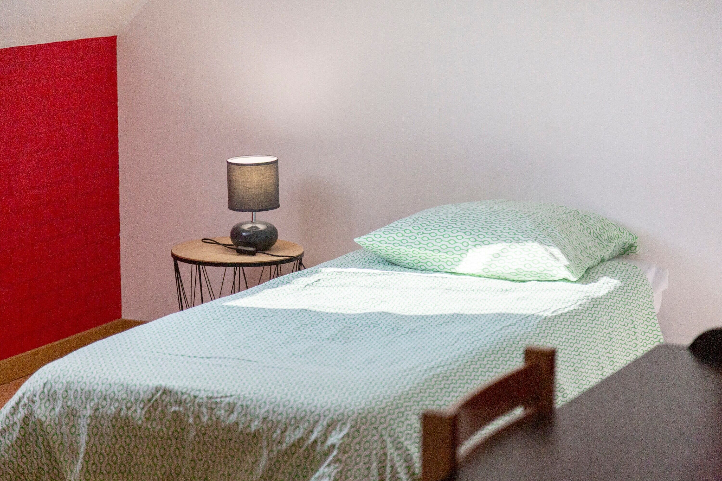 Un lit simple avec une couvre-lit à motifs, une lampe de chevet et une petite table ronde avec une lampe.