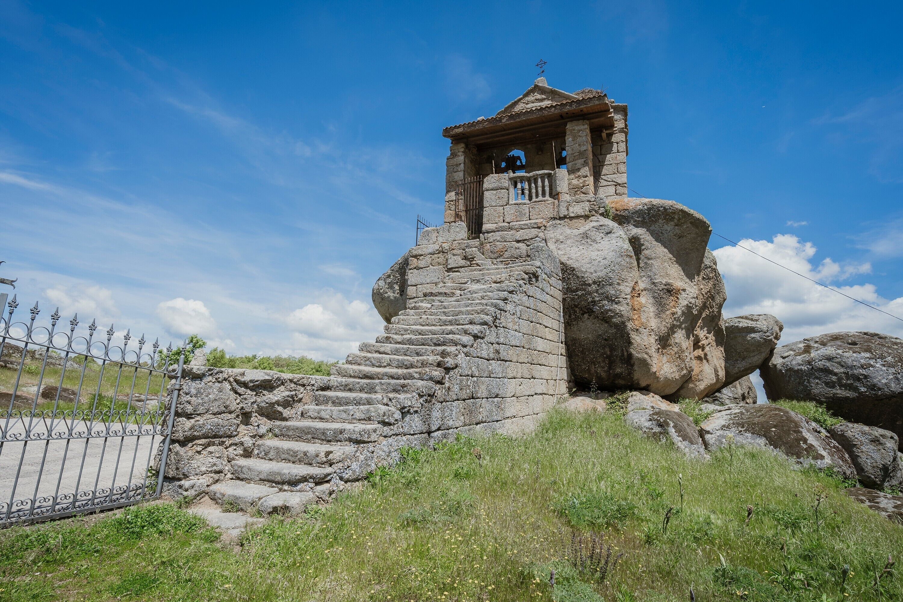 Une structure en pierre avec un escalier menant à un petit bâtiment situé en haut, entourée de gros rochers et d’une clôture métallique.