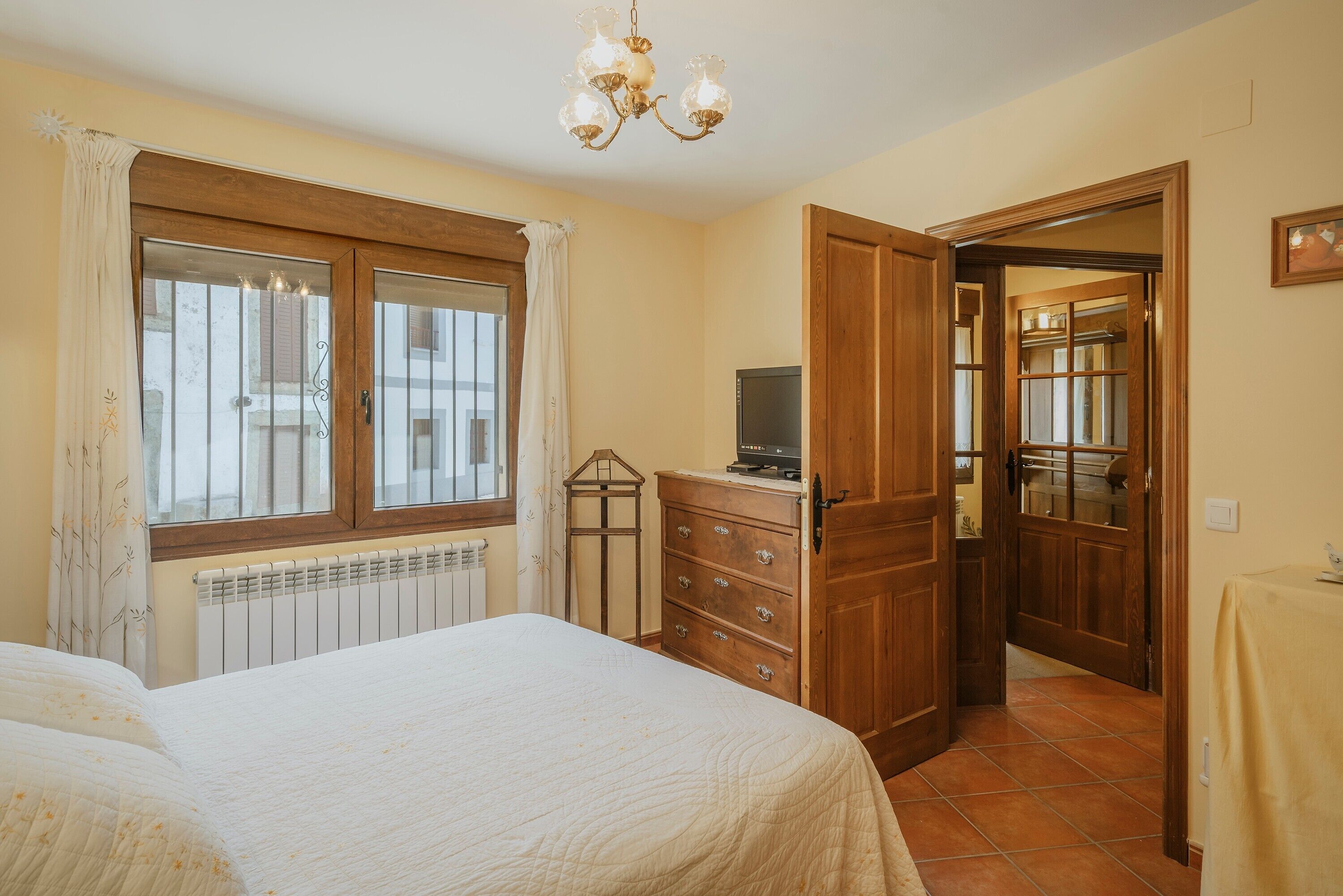 A bedroom with a bed, a wooden dresser, a radiator, a television, and a chandelier.