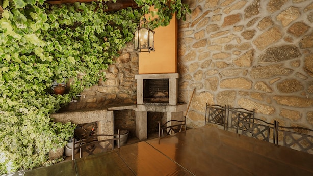 Pared de piedra con chimenea, una mesa con sillas y follaje verde.