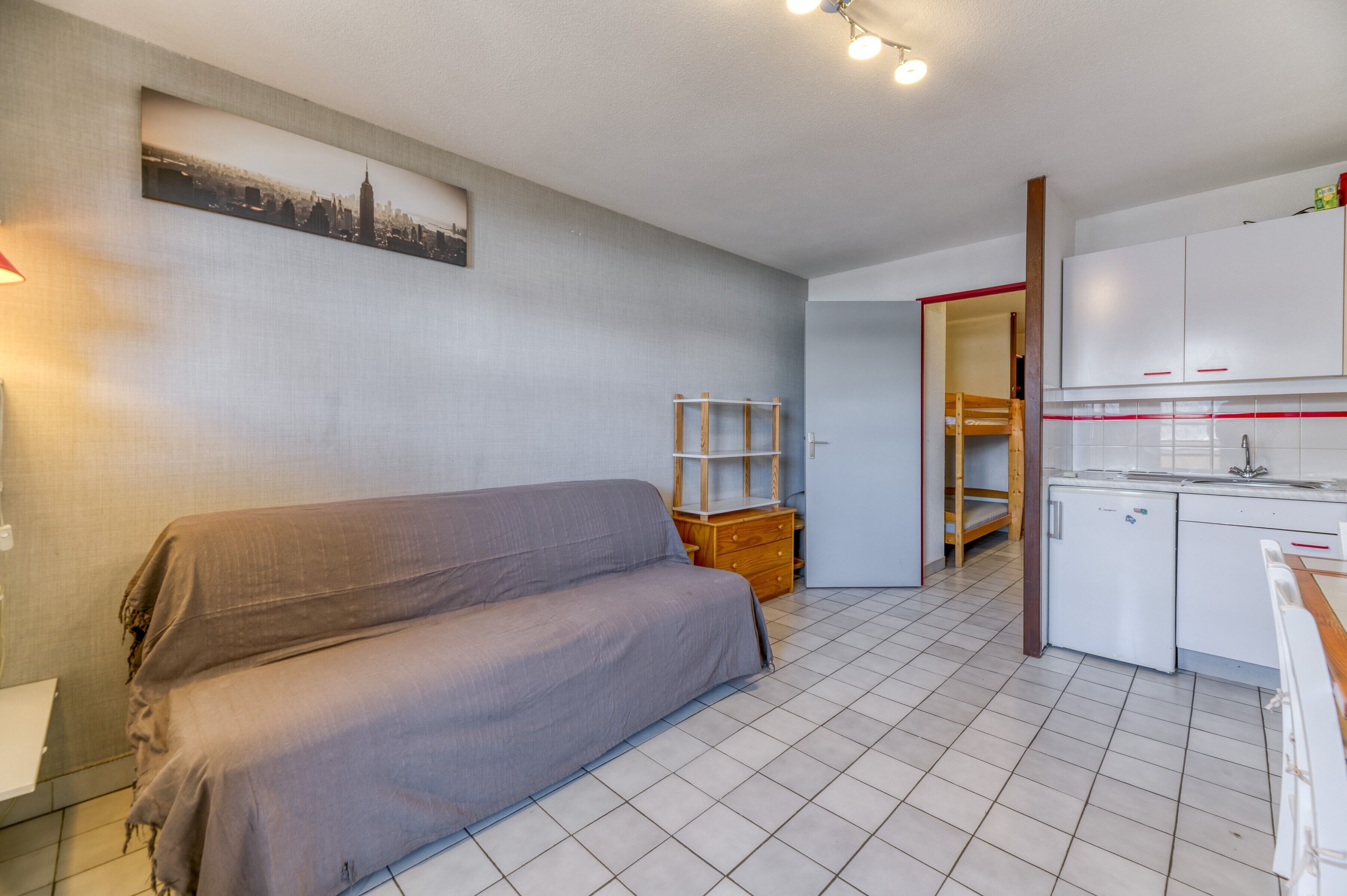Un espace de vie compact comprenant un canapé, un petit meuble en bois, une kitchenette et un tableau représentant un paysage urbain accroché au mur.
