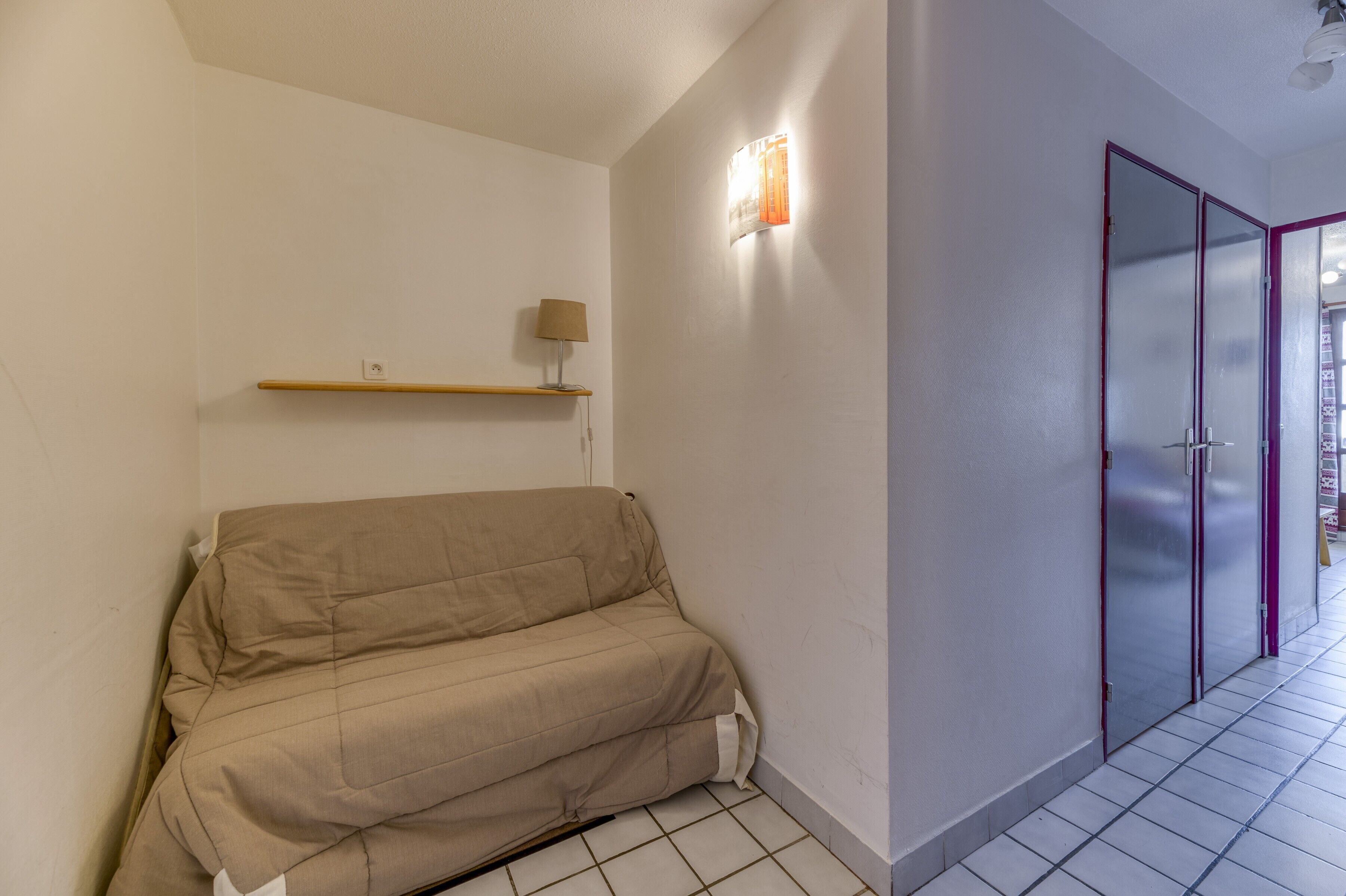 Une pièce avec un canapé beige, une étagère sur laquelle se trouve une lampe, et une porte dont les panneaux sont en verre dépoli.