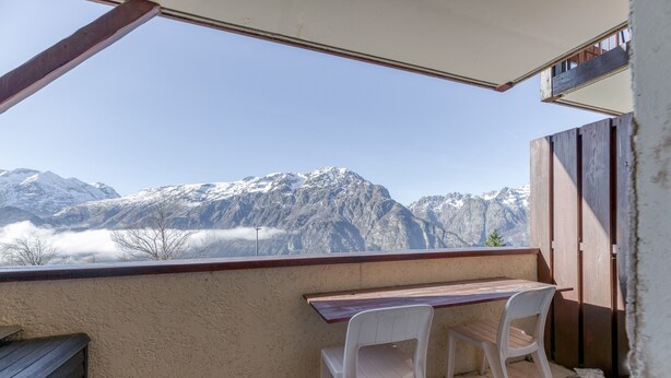Un balcón con una mesa de madera y dos sillas blancas de plástico, con vistas a montañas nevadas.
