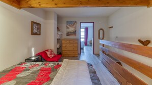 Ein Schlafzimmer mit Holzboden, einem Bett mit Decke, einem Schminktisch und einem Spiegel, sowie Blick in einen anderen Raum.
