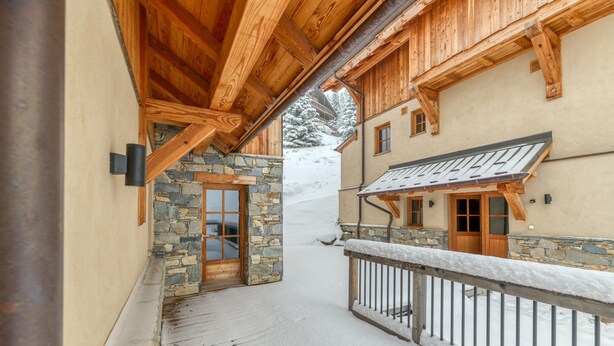 Un sendero cubierto de nieve que conduce a una edificación de madera con detalles de piedra y un balcón con barandilla de metal.